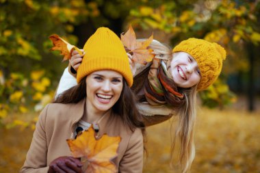 Hello november. smiling modern mother and child in orange hats with autumn yellow leaves having fun time outdoors in the city park in autumn.