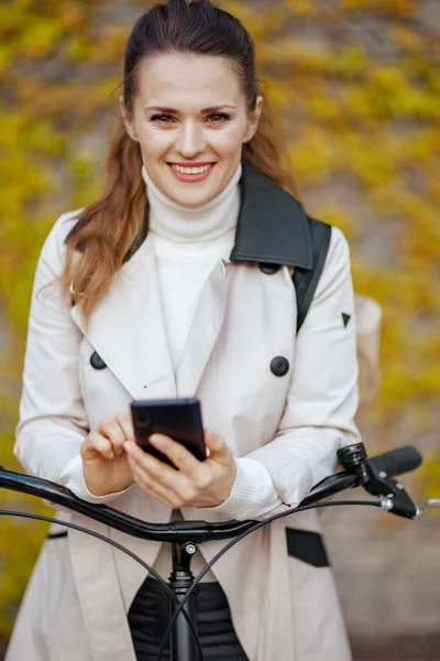 Portrait of happy modern woman in beige trench coat with bicycle using smartphone app against the green wall outside in the city.
