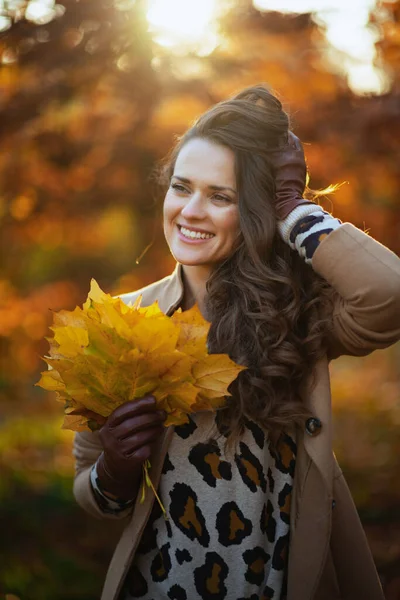 Hello september. happy 40 years old woman in beige coat with autumn yellow leaves outside in the city park in autumn.