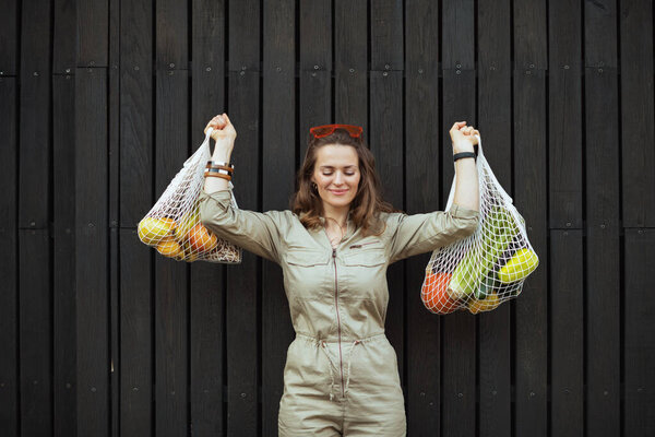 Улыбающаяся модная женщина в целом со струнной сумкой против черной деревянной стены на открытом воздухе в городе.