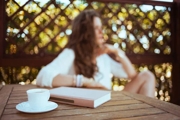 Closeup on cup of coffee and book at the table and woman in background in the terrace.