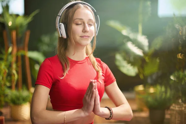 modern woman in red fitness clothes with headphones meditating at modern green home.