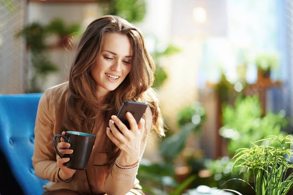 Green Home. smiling stylish middle aged woman with long wavy hair with cup of cappuccino using smartphone app in the modern living room in sunny day.