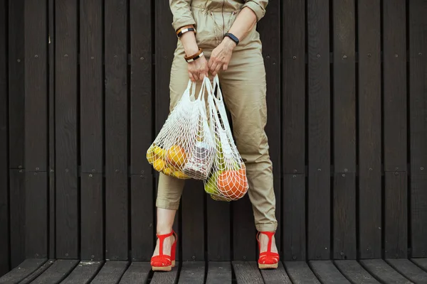 Closeup on woman in overall with string bag against black wood wall outside in the city.