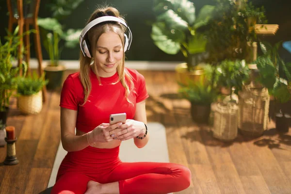 modern woman in red fitness clothes with smartphone listening to the music with headphones in the modern green living room.