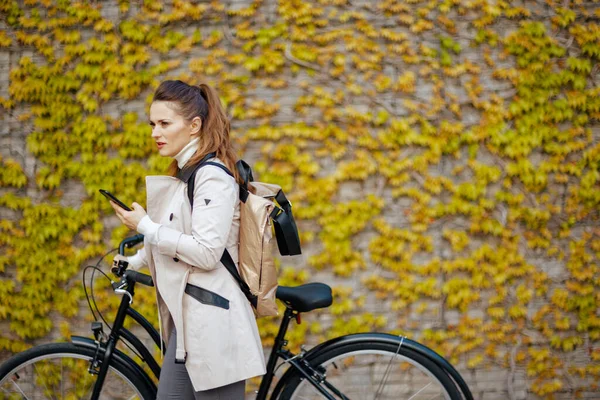 Pensive Stylish Female Beige Trench Coat Bicycle Backpack Using Smartphone Stockbild