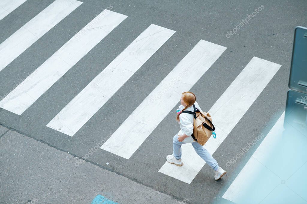 Upper view of modern girl in white sweatshirt with backpack crossing crosswalk and going to school outdoors in the city.