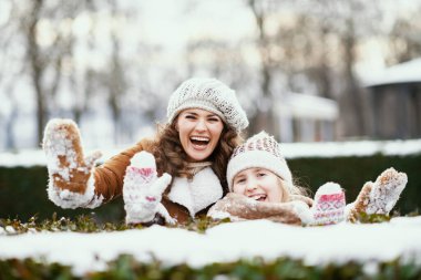 Örgü şapkalar ve koyun derisi ceketlerle mutlu bir anne ve kız kışın şehir parkında kar eldivenlerini gösteren örülü bir şapka ve koyun derisi bir paltoyla..
