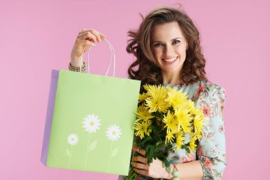 Portrait of smiling young female in floral dress with yellow chrysanthemums flowers and green shopping bag isolated on pink.