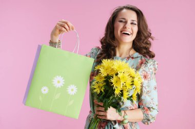 happy modern female with long wavy brunette hair with yellow chrysanthemums flowers and green shopping bag isolated on pink background.