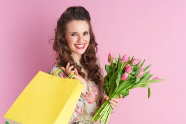 happy young woman with long wavy brunette hair with tulips bouquet and shopping bags isolated on pink.