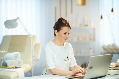 smiling 40 years old woman worker with laptop working in modern beauty studio.