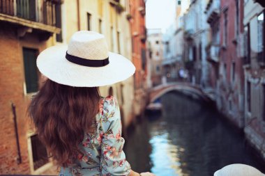 Seen from behind modern woman in floral dress with hat enjoying promenade in Venice, Italy.