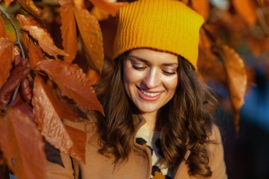 Hello autumn. smiling elegant woman in brown coat and yellow hat among autumnal foliage outside on the city park in autumn.