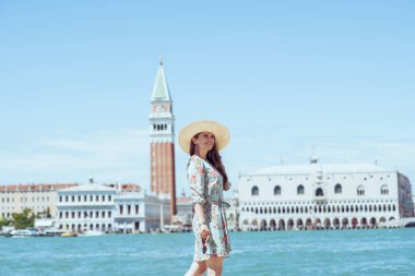 Gülümseyen orta yaşlı, çiçekli elbiseli, güneş gözlüklü, şapkalı, San Giorgio Maggiore adasında yürüyüş yapan bir gezgin kadın..