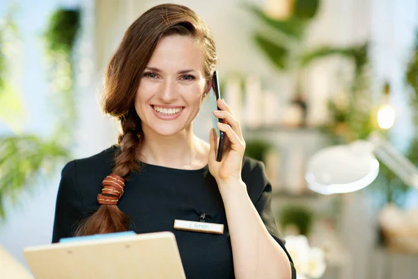 smiling 40 years old woman employee with clipboard speaking on a smartphone in modern beauty salon.