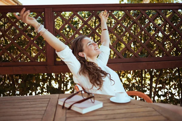 happy modern woman in white shirt with cup of coffee, book and eyeglasses sitting at the table in the patio of guest house hotel with raised arms rejoicing.