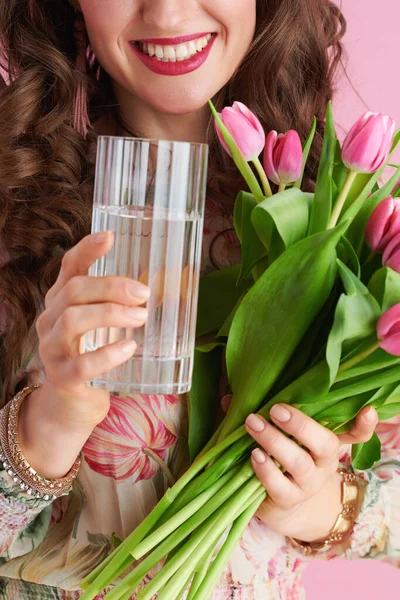 Closeup on happy woman with tulips bouquet and glass of water isolated on pink background.