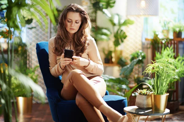 Green Home. stylish middle aged housewife with long wavy hair at modern home in sunny day in beige pants and blouse sending text message using smartphone while sitting in a blue armchair.