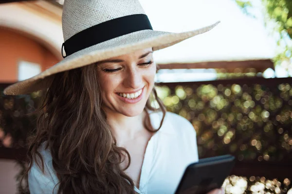 smiling trendy woman in white shirt with hat using smartphone applications in the patio of guest house hotel.