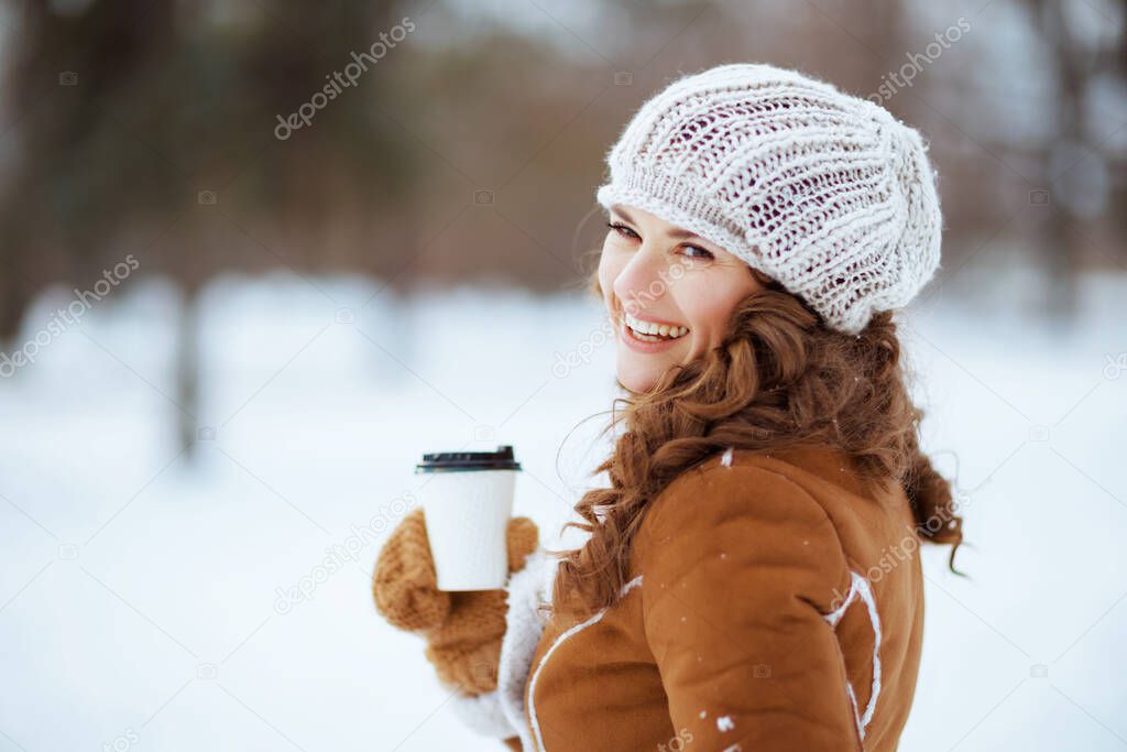 smiling stylish middle aged woman with mittens and cup of hot cocoa in a knitted hat and sheepskin coat outdoors in the city park in winter.