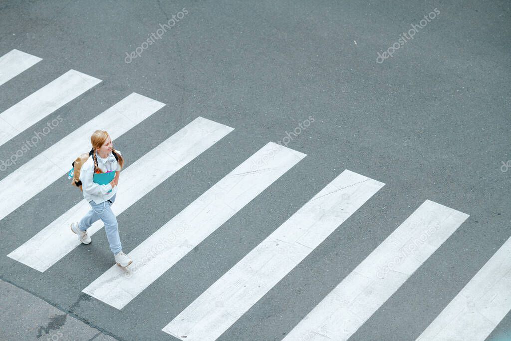 Upper view of smiling trendy pupil in white sweatshirt with workbook, backpack and headphones crossing crosswalk and going to school outdoors in the city.