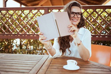 happy stylish middle aged housewife in white shirt with cup of coffee, book and eyeglasses sitting at the table in the patio.