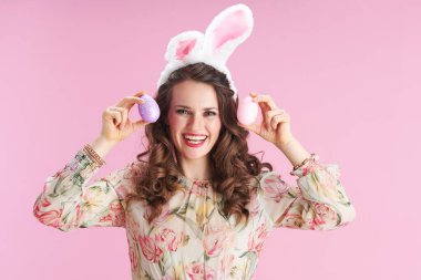 Uzun dalgalı esmer saçlı, tavşan kulaklı ve pembe arka planda paskalya yumurtası olan genç bir kadın..