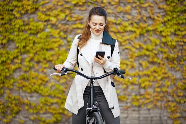 smiling modern woman in beige trench coat with bicycle sending text message using smartphone against the green wall outdoors on the city street.