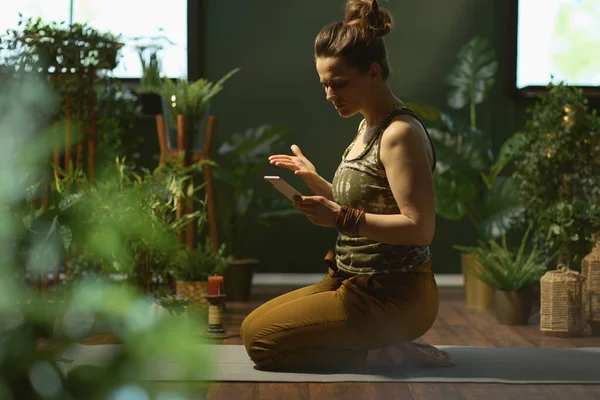 sad modern woman with smartphone doing yoga at modern green home.