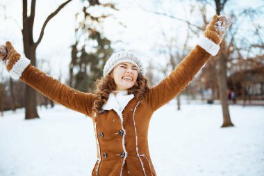 Örgü şapkalı ve koyun derisi ceketli mutlu orta yaşlı kadın kışın şehir parkında neşe içinde eğleniyor..