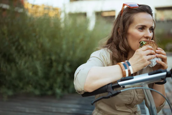 elegant middle aged woman in overall with scooter and sandwich eating while sitting outdoors in the city.