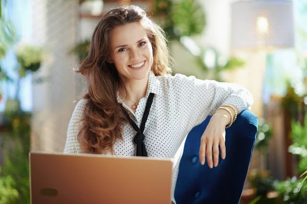 Green Home. smiling trendy woman with long wavy hair with laptop in the modern living room in sunny day.