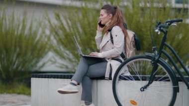 smiling elegant female in beige trench coat with bicycle and laptop speaking on a smartphone while sitting on the bench outdoors in the city.