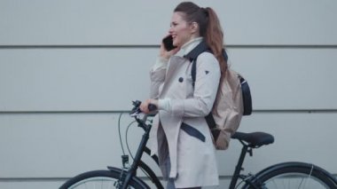 modern female in beige trench coat with bicycle and backpack using a smartphone In front of a metal wall outdoors in the city.