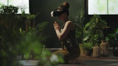 stylish 40 years old housewife with VR headset doing yoga in metaverse in the modern green living room.