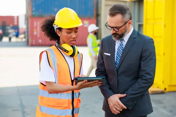 Caucasian business man and black female dock worker look at digital tablet computer working at warehouse container yard.
