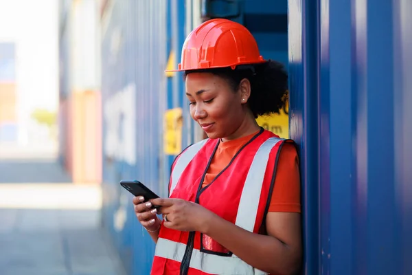 Online working by smartphone. Black female dock worker control loading containers box from cargo at warehouse container yard. Marine and carrier insurance concept. logistic shipping yard