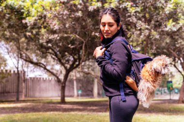 A charming young woman walks in nature, with a backpack on her shoulder, from which her dog peeks out.