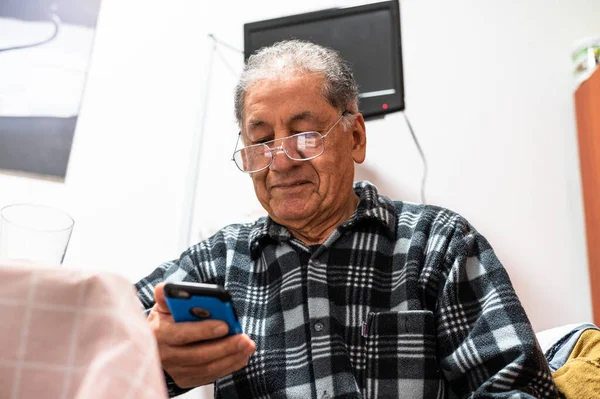 Homem caucasiano sênior feliz em óculos olhar para tela do celular navegar internet sem fio. Sorrindo moderno maduro 70 texto do avô ou mensagem no smartphone. Idosos usam tecnologias celulares em casa. Imagem De Stock