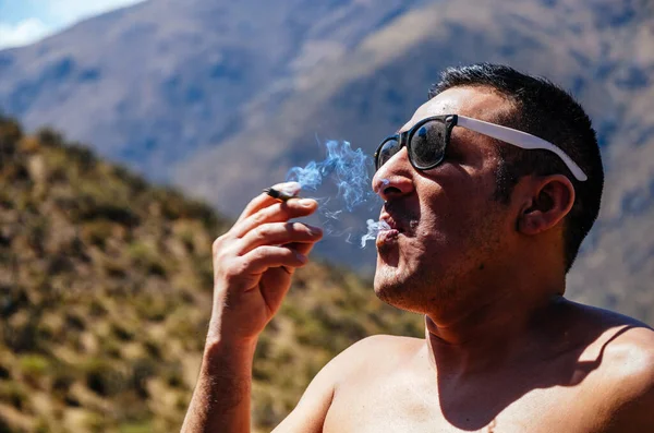 Jovem fumando tabaco, homem com óculos de sol e fundo de montanhas . Fotos De Bancos De Imagens