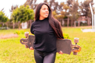 Elinde kaykayıyla gülümseyen genç bayan kaykaycının portresi. Parkta kaykay yapan bir kadın.