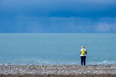 Sırt çantalı yalnız kadın denizin kenarında duruyor. Bulutlu gökyüzü ve soğuk su. Yağmur geliyor.