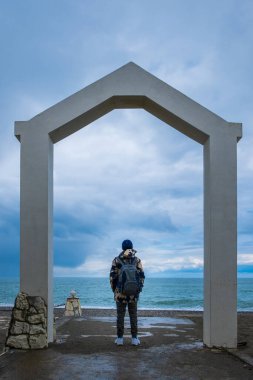 Şapkalı ve sırt çantalı bir adam kemerin altında duruyor ve denize bakıyor. Deniz ufku ve gökyüzü. Deniz kıyısında gezgin