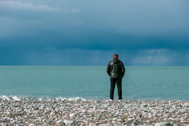 Deri ceketli bir adam kıyıda duruyor ve denize bakıyor. Gökyüzü bulutlu. Deniz kenarında yalnızlık