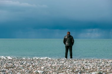 Deri ceketli bir adam kıyıda duruyor ve denize bakıyor. Gökyüzü bulutlu. Deniz kenarında yalnızlık