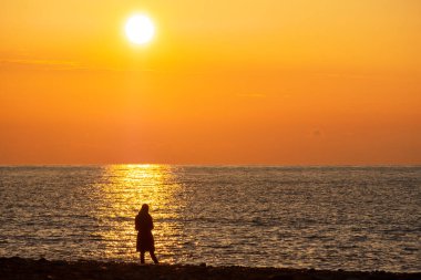 Gün batımına karşı duran bir adamın silueti. Denizin arka planında bir adam. Akşam, turuncu gökyüzüne doğru yürü.