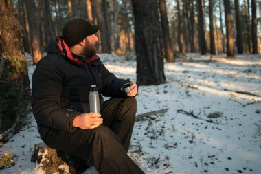 Sakallı, orta yaşlı bir adam termosla ormanda oturur ve çay içer. Ormana bakıyorum.