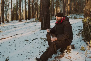 Yetişkin sakallı bir adam ormanda elinde bir kupa çay ve termosla oturuyor. Onun önünde düşünceli görünüyor.