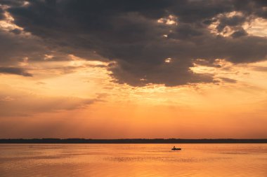 Yaz günü gün batımında göl manzarası. Gölde kayıktaki bir adamın silüeti. Akşamüstü göl turuncu renkli.